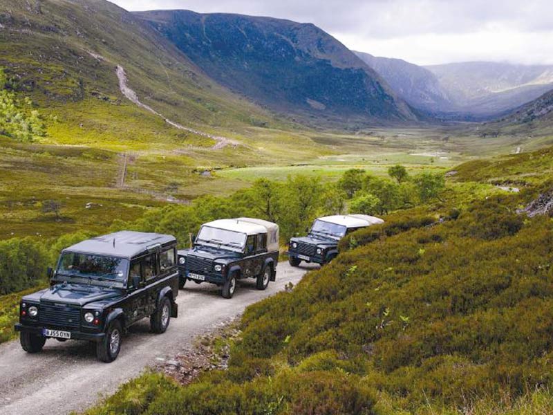 Three Land Rover Defenders driving through a misty Highland mountain landscape on a gravel road.